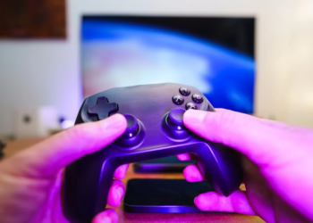 Hands holding a black game controller in front of a blurry TV showing abstract blue light, pink ambient lighting