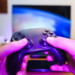 Hands holding a black game controller in front of a blurry TV showing abstract blue light, pink ambient lighting