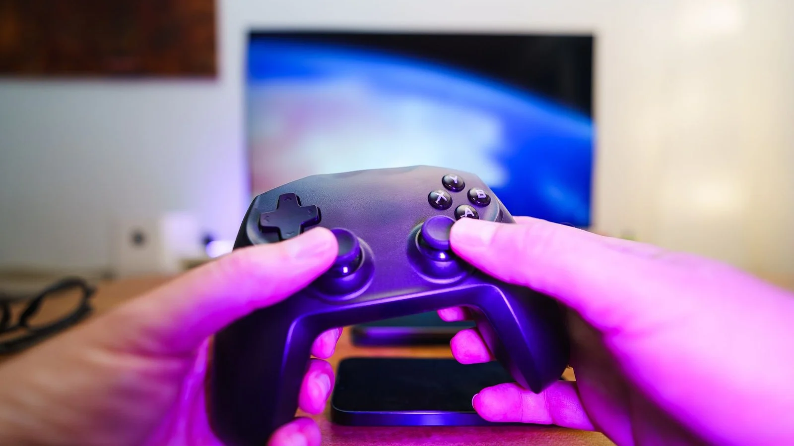 Hands holding a black game controller in front of a blurry TV showing abstract blue light, pink ambient lighting
