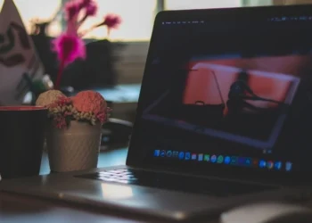 An open MacBook on a desk showing the macOS desktop and dock, beside a mug and flowers