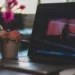 An open MacBook on a desk showing the macOS desktop and dock, beside a mug and flowers