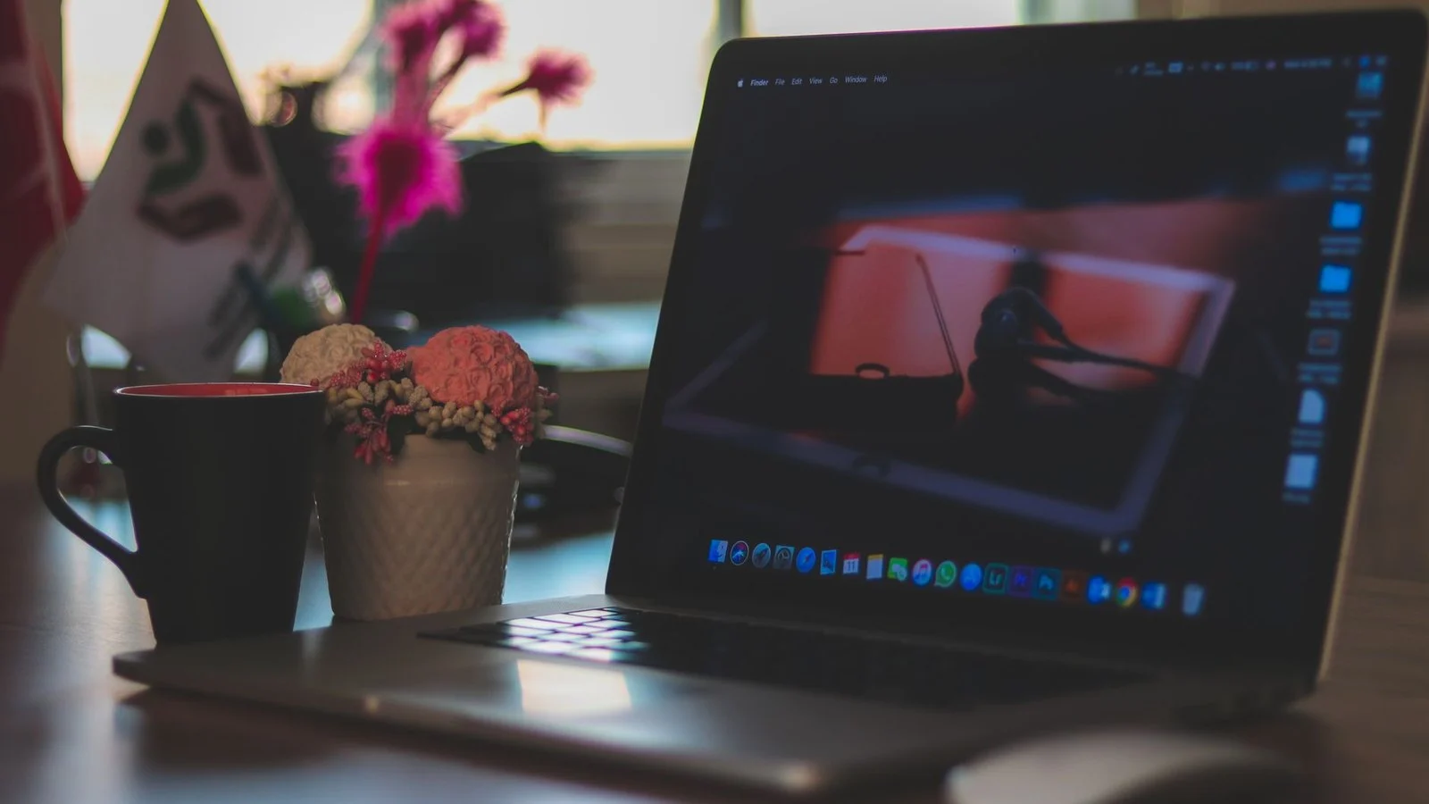 An open MacBook on a desk showing the macOS desktop and dock, beside a mug and flowers