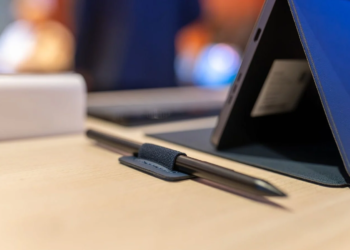 A dark stylus tucked in a fabric loop beside a tablet in a blue folio case on a wooden desk