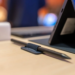 A dark stylus tucked in a fabric loop beside a tablet in a blue folio case on a wooden desk