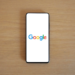 Black-bezeled smartphone lying flat on wood desk, screen showing the Google logo