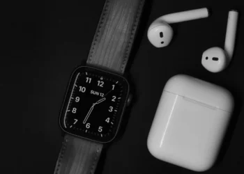 black square smartwatch with analog numeral face and dark strap beside white earbuds and charging case