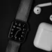 black square smartwatch with analog numeral face and dark strap beside white earbuds and charging case