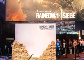 Crowd queues at a Rainbow Six Siege gamescom booth beneath a large game banner and broken brick wall prop