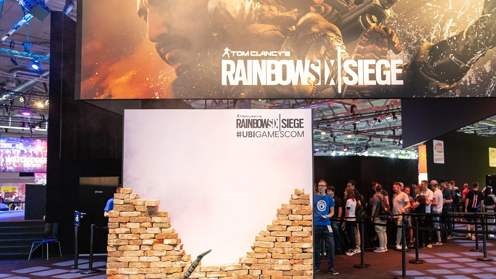 Crowd queues at a Rainbow Six Siege gamescom booth beneath a large game banner and broken brick wall prop
