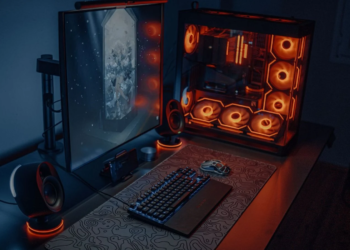 A dark gaming room with an open-panel desktop PC lit by orange RGB fans beside a monitor, mechanical keyboard and mouse