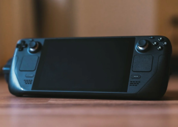 A black handheld gaming console with dual grips and joysticks resting on a wooden table, screen off