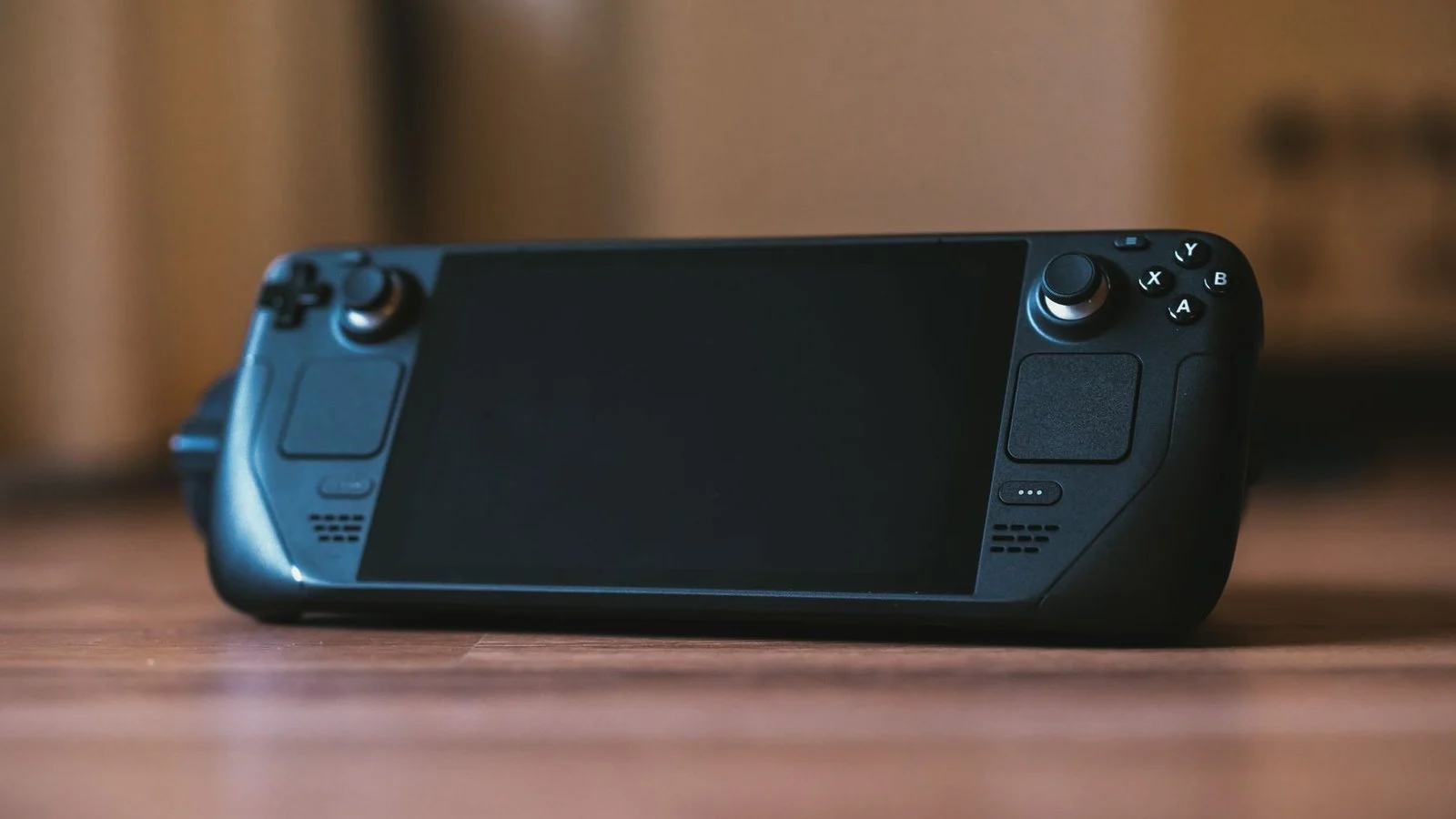 A black handheld gaming console with dual grips and joysticks resting on a wooden table, screen off