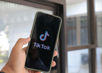 Hand holding a smartphone displaying the TikTok logo and splash screen near a window