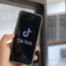 Hand holding a smartphone displaying the TikTok logo and splash screen near a window