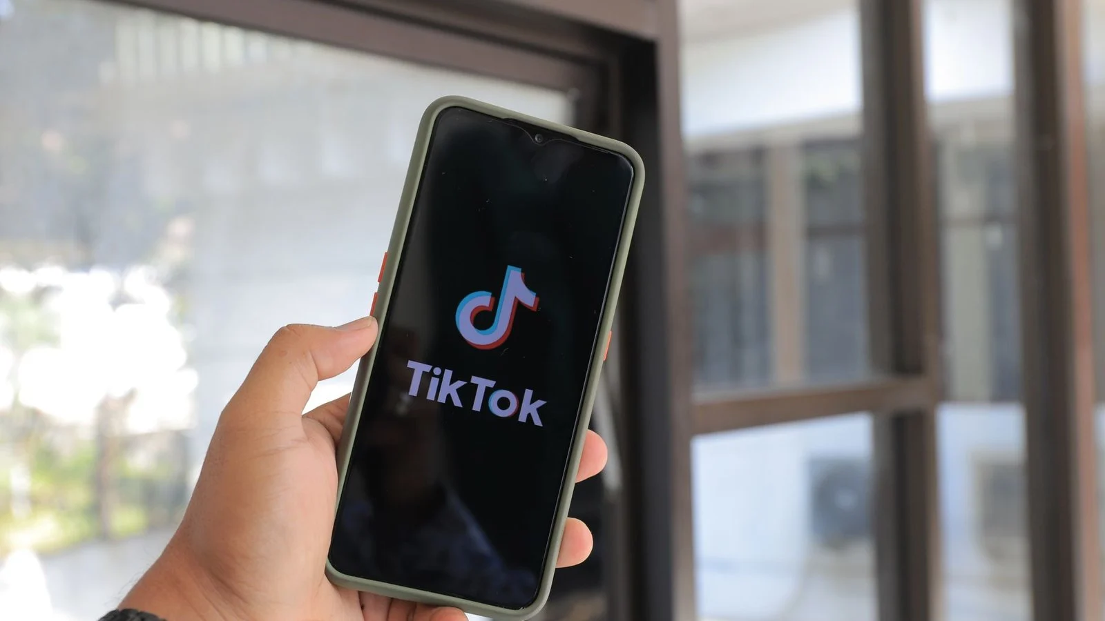 Hand holding a smartphone displaying the TikTok logo and splash screen near a window