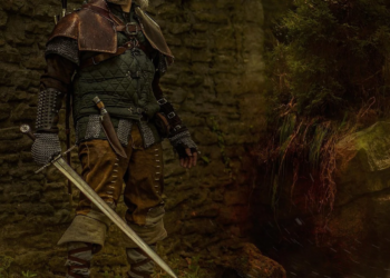 Person in medieval-style leather and chainmail armor holding a longsword near a stone ruin