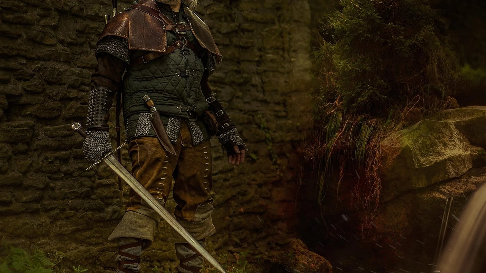 Person in medieval-style leather and chainmail armor holding a longsword near a stone ruin