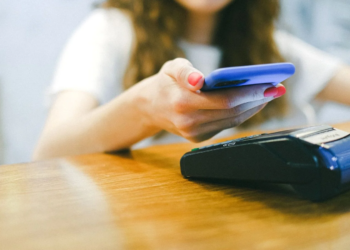 Woman taps a phone in a blue case against a wireless card payment terminal on a wooden counter