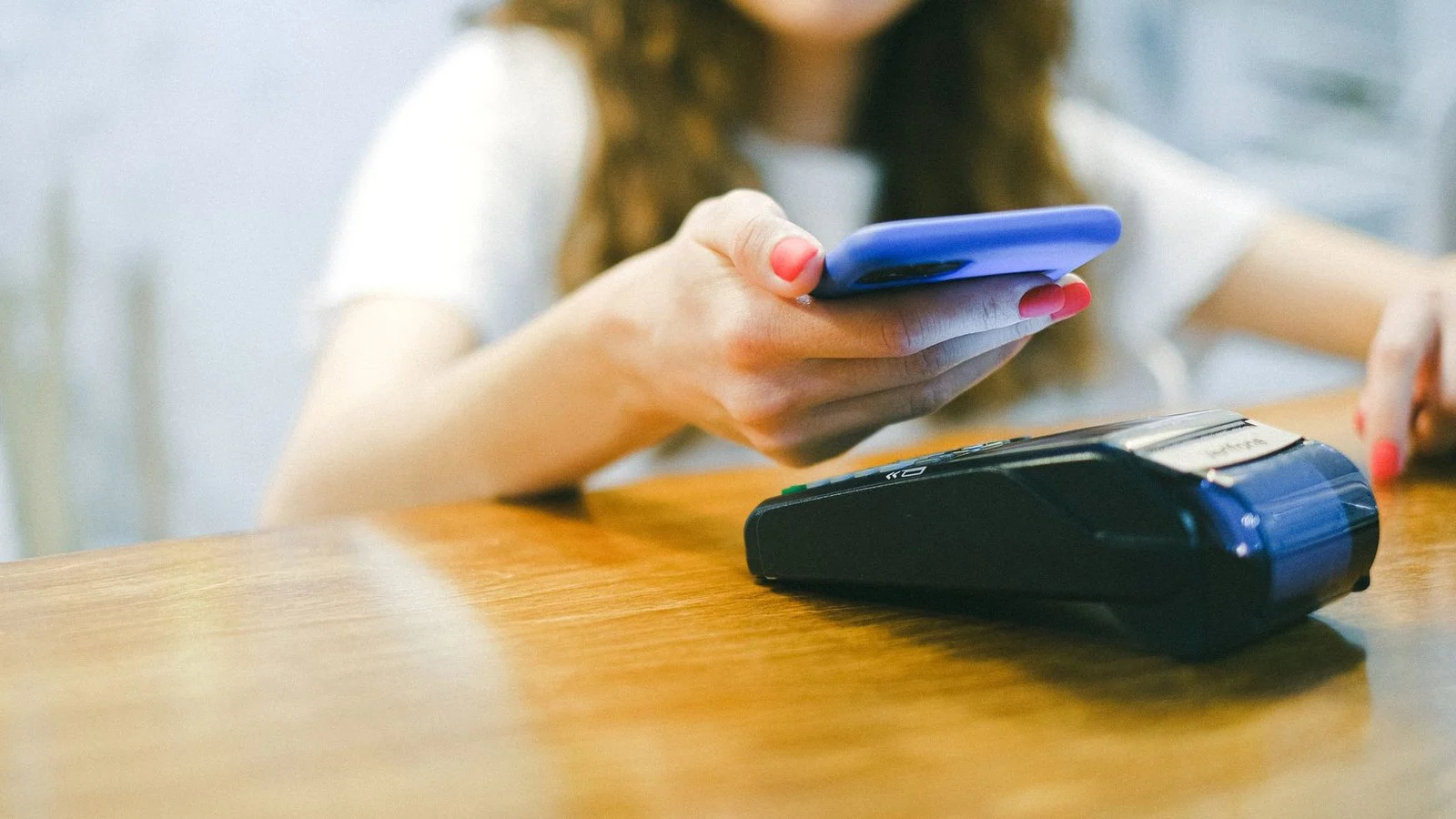 Woman taps a phone in a blue case against a wireless card payment terminal on a wooden counter