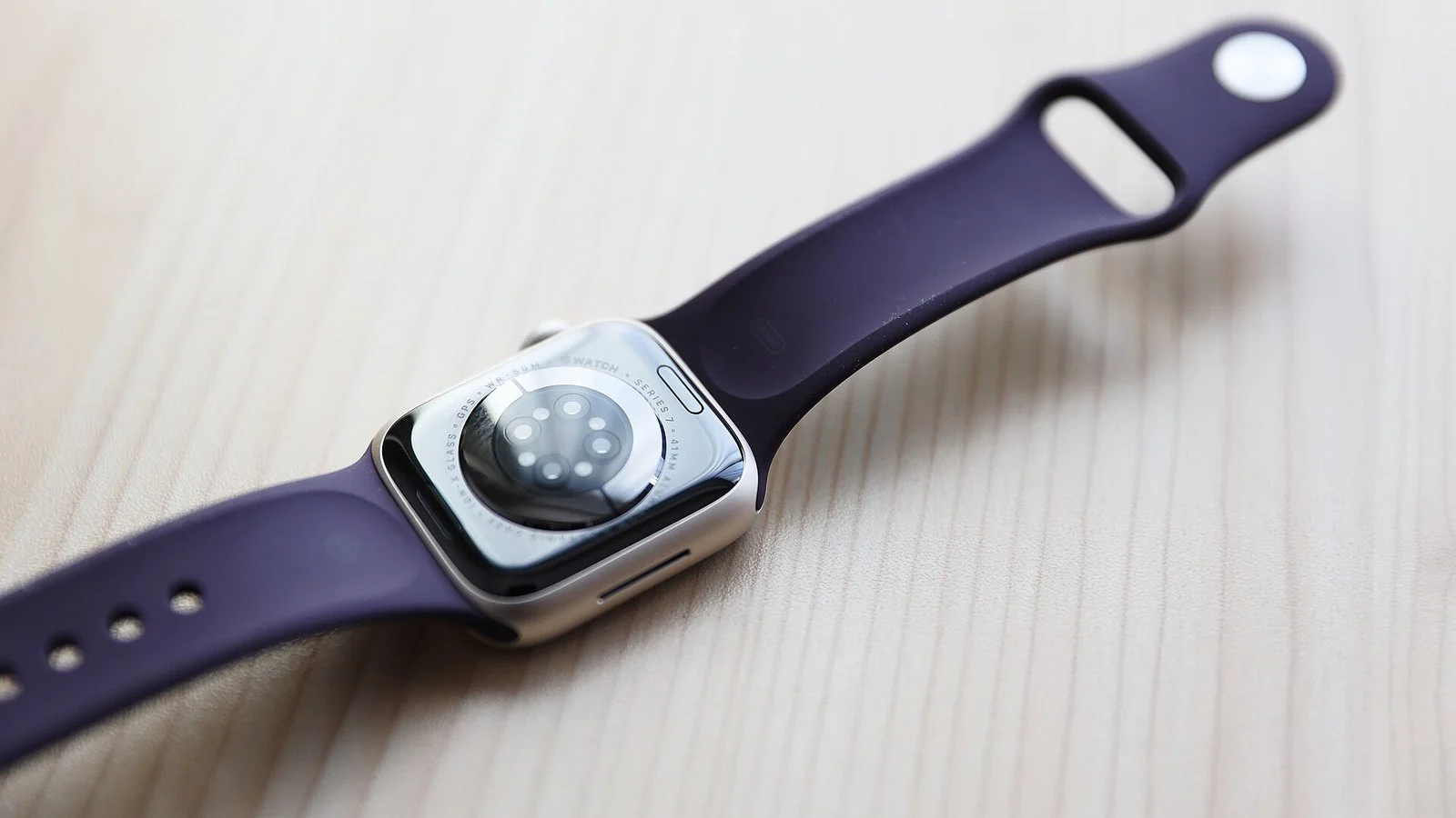 Apple Watch Series 7 with purple silicone band, face-down showing sensor cluster on a wood surface