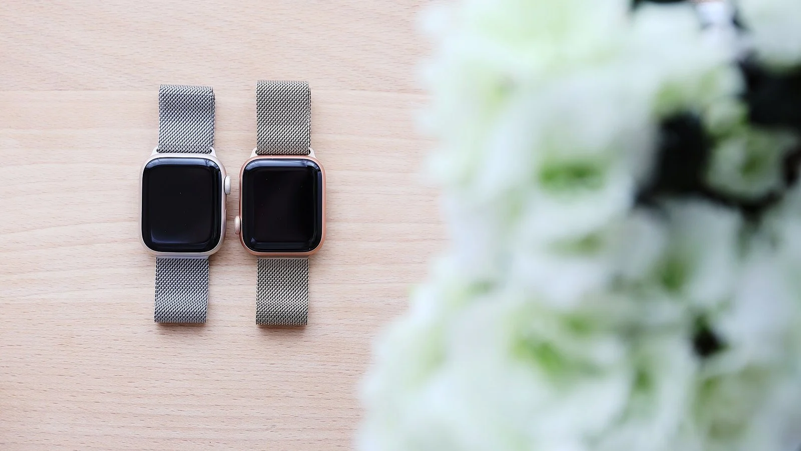 Two Apple Watches with metal mesh bands and dark screens lie on a wooden surface