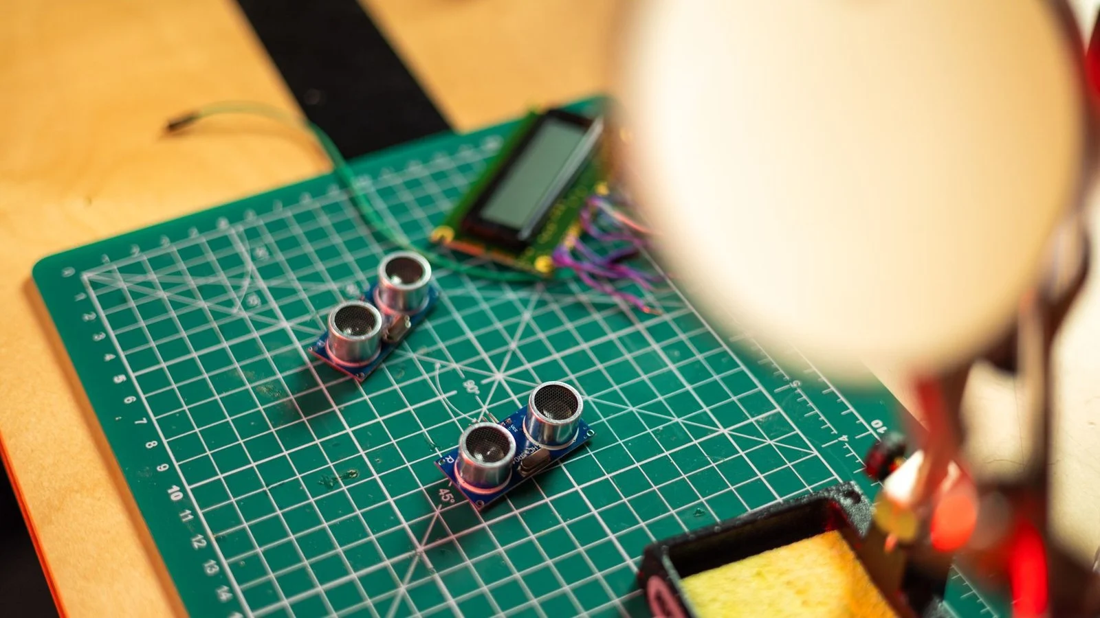 Ultrasonic sensor modules and a small LCD on a green cutting mat near a soldering iron
