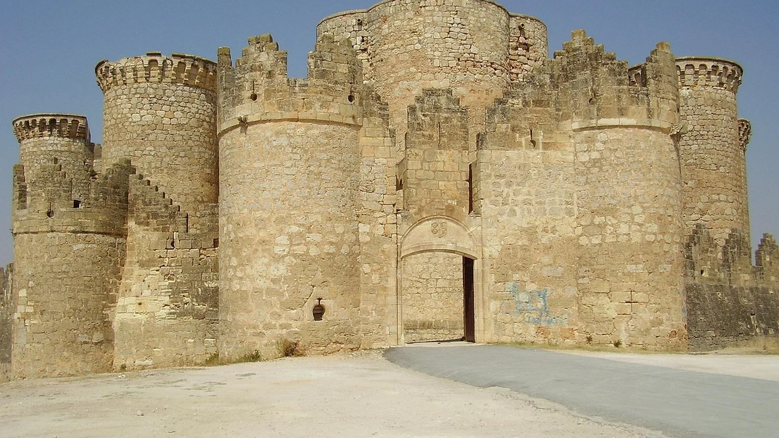 Stone medieval castle with crenellated towers and an arched gate, blue sky, graffiti on the wall