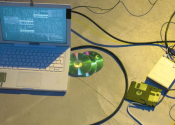 Laptop showing terminal text beside a floppy disk, floppy drive, and CDs, with cables on a concrete floor