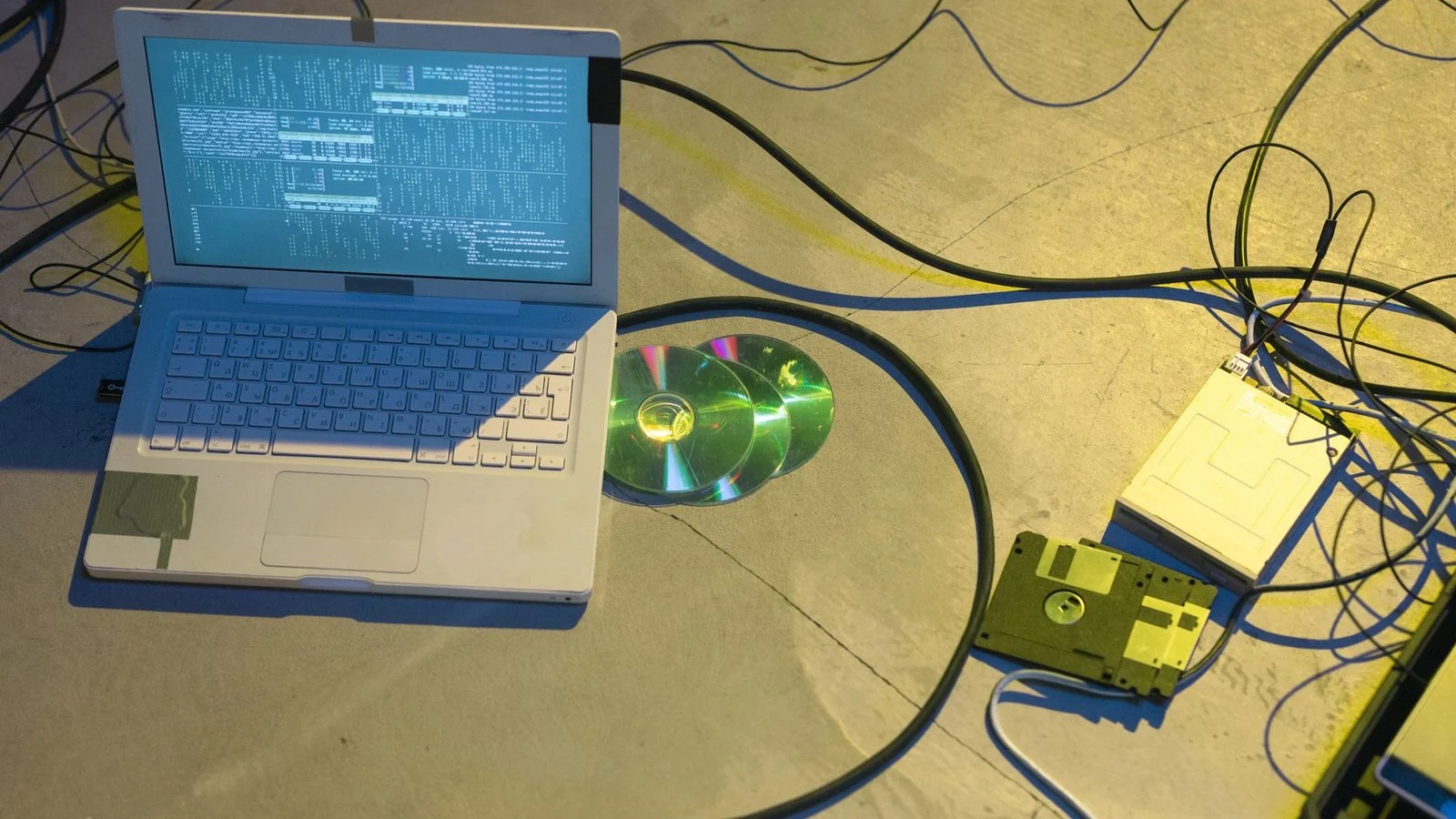Laptop showing terminal text beside a floppy disk, floppy drive, and CDs, with cables on a concrete floor