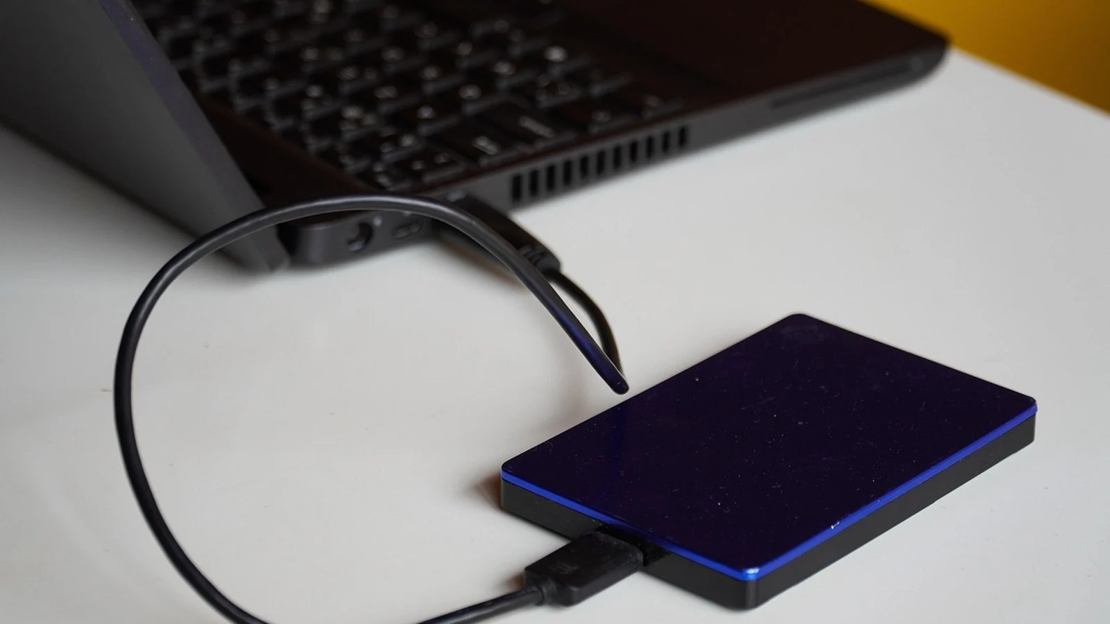 A blue and black portable external hard drive connected by USB cable to a laptop on a white desk