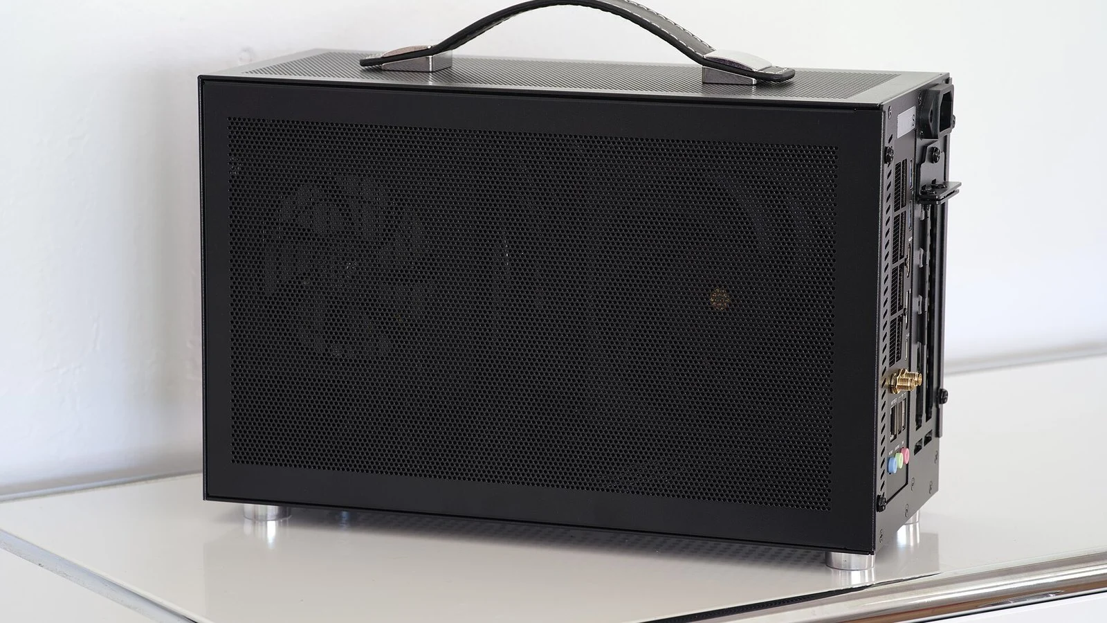 Black mesh-paneled mini-PC with leather carry handle on a white table, side panel closed