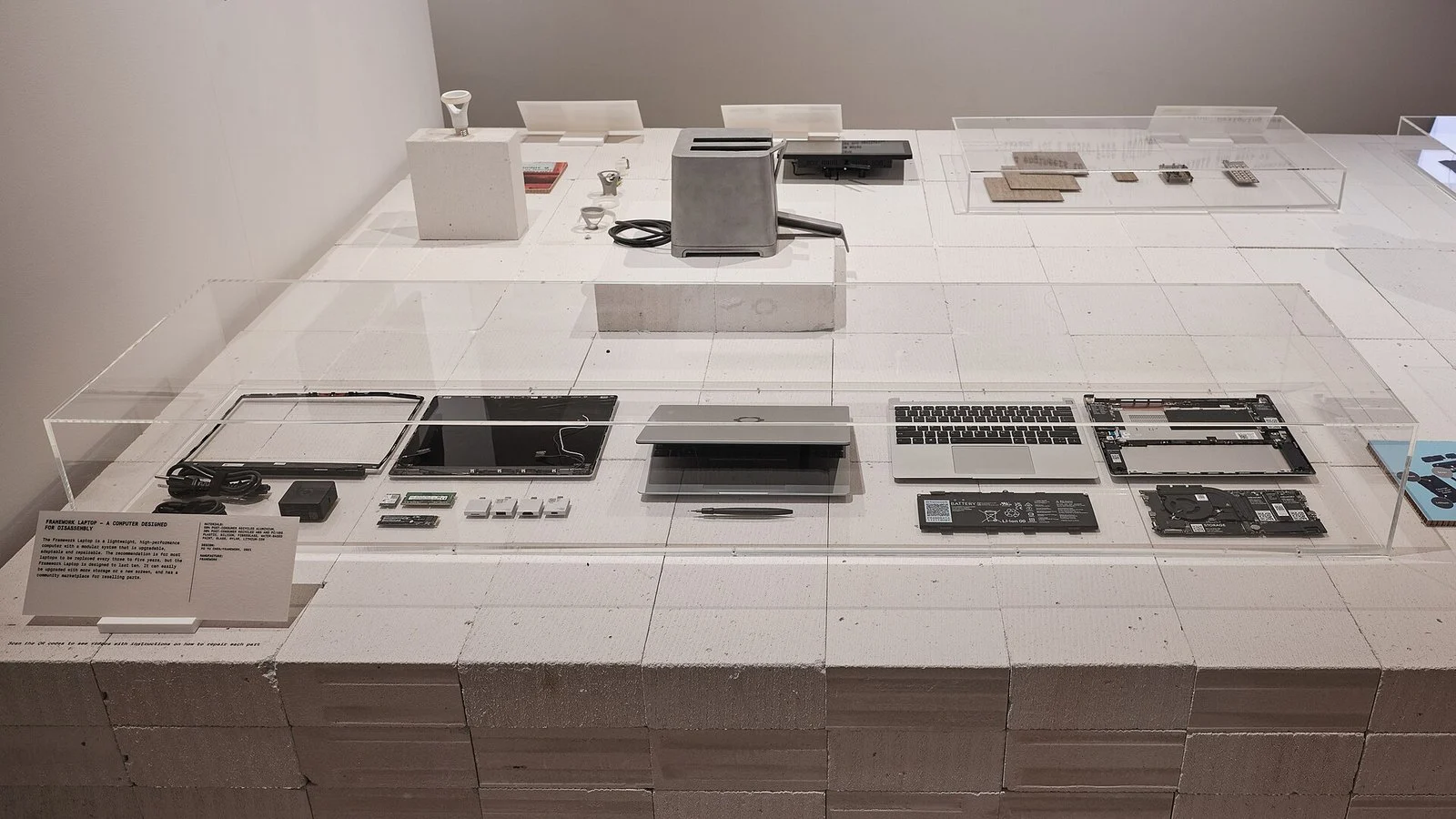 Disassembled Framework Laptop parts displayed in a museum exhibit case on pricing repairability