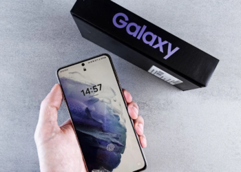 Hand holding a Samsung Galaxy phone with lock screen showing, box labeled Galaxy behind it