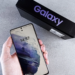 Hand holding a Samsung Galaxy phone with lock screen showing, box labeled Galaxy behind it