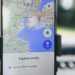 Smartphone in a car dashboard mount showing Google Maps overview of New York with an Explore Nearby panel