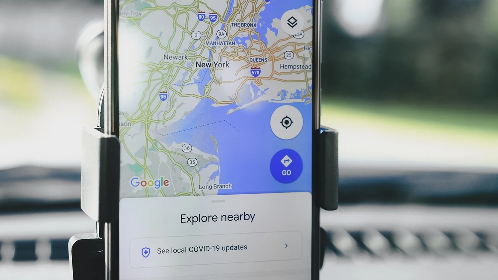 Smartphone in a car dashboard mount showing Google Maps overview of New York with an Explore Nearby panel