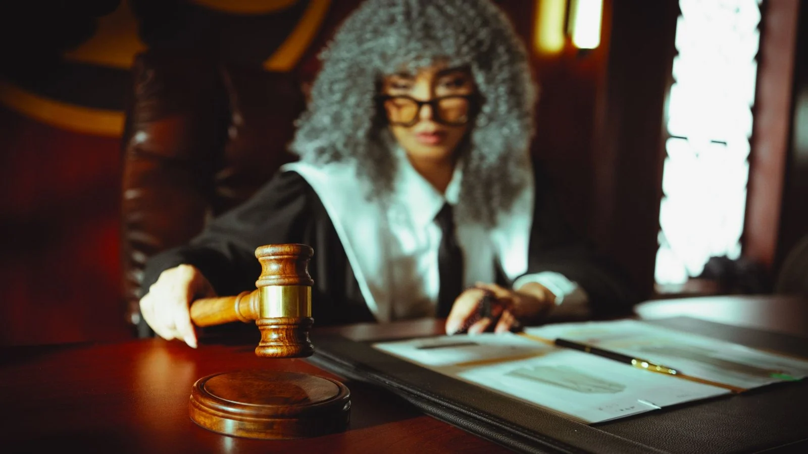 A judge in robes and a curly grey wig holds a gavel beside papers on a courtroom bench
