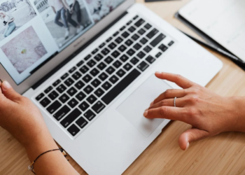 Hands on a silver laptop trackpad and keyboard, screen showing a grid of photo thumbnails