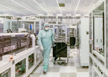 Worker in cleanroom suit pushes equipment cart through a semiconductor fab aisle