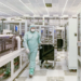 Worker in cleanroom suit pushes equipment cart through a semiconductor fab aisle