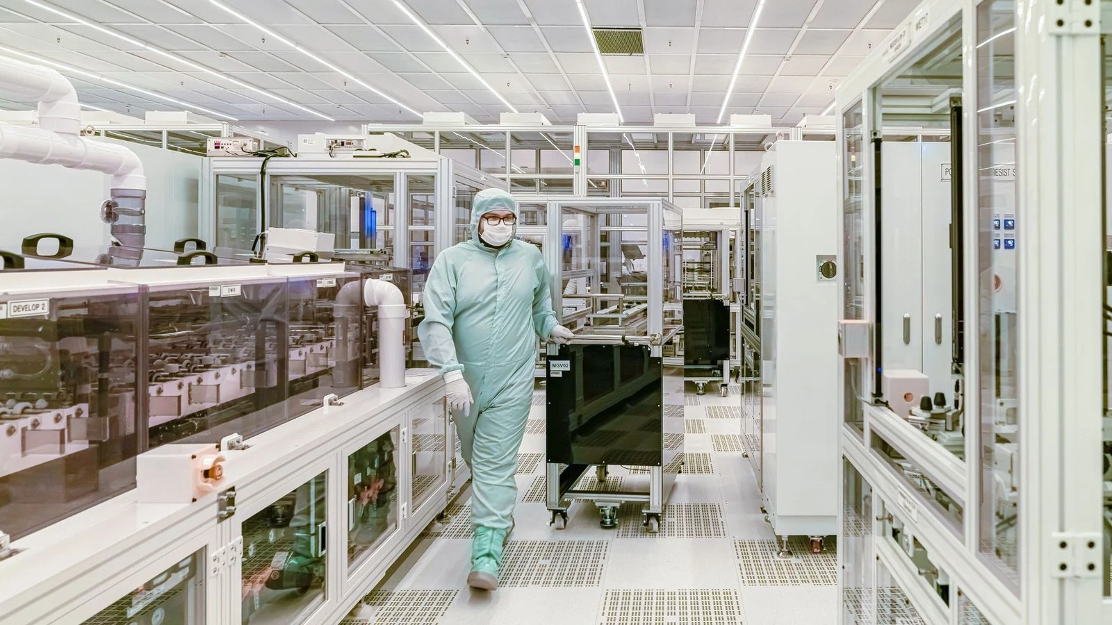 Worker in cleanroom suit pushes equipment cart through a semiconductor fab aisle