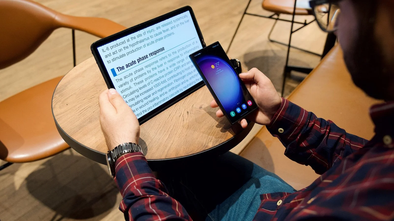 Man holds a Samsung phone while reading a medical text article on a flat tablet at a cafe table