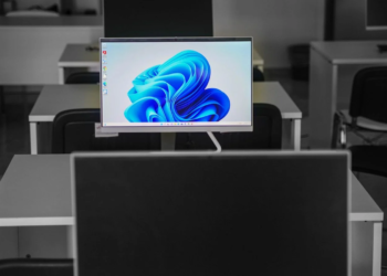 A slim all-in-one desktop PC showing the Windows 11 wallpaper among empty desks in a computer room