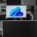 A slim all-in-one desktop PC showing the Windows 11 wallpaper among empty desks in a computer room