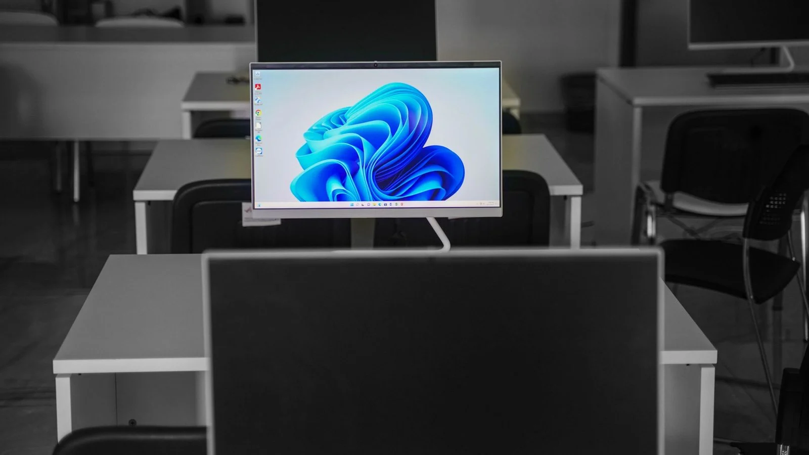 A slim all-in-one desktop PC showing the Windows 11 wallpaper among empty desks in a computer room