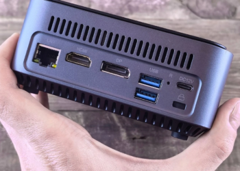 Hand holding a small gray mini PC showing LAN, HDMI, DP, USB, and power ports