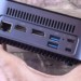 Hand holding a small gray mini PC showing LAN, HDMI, DP, USB, and power ports