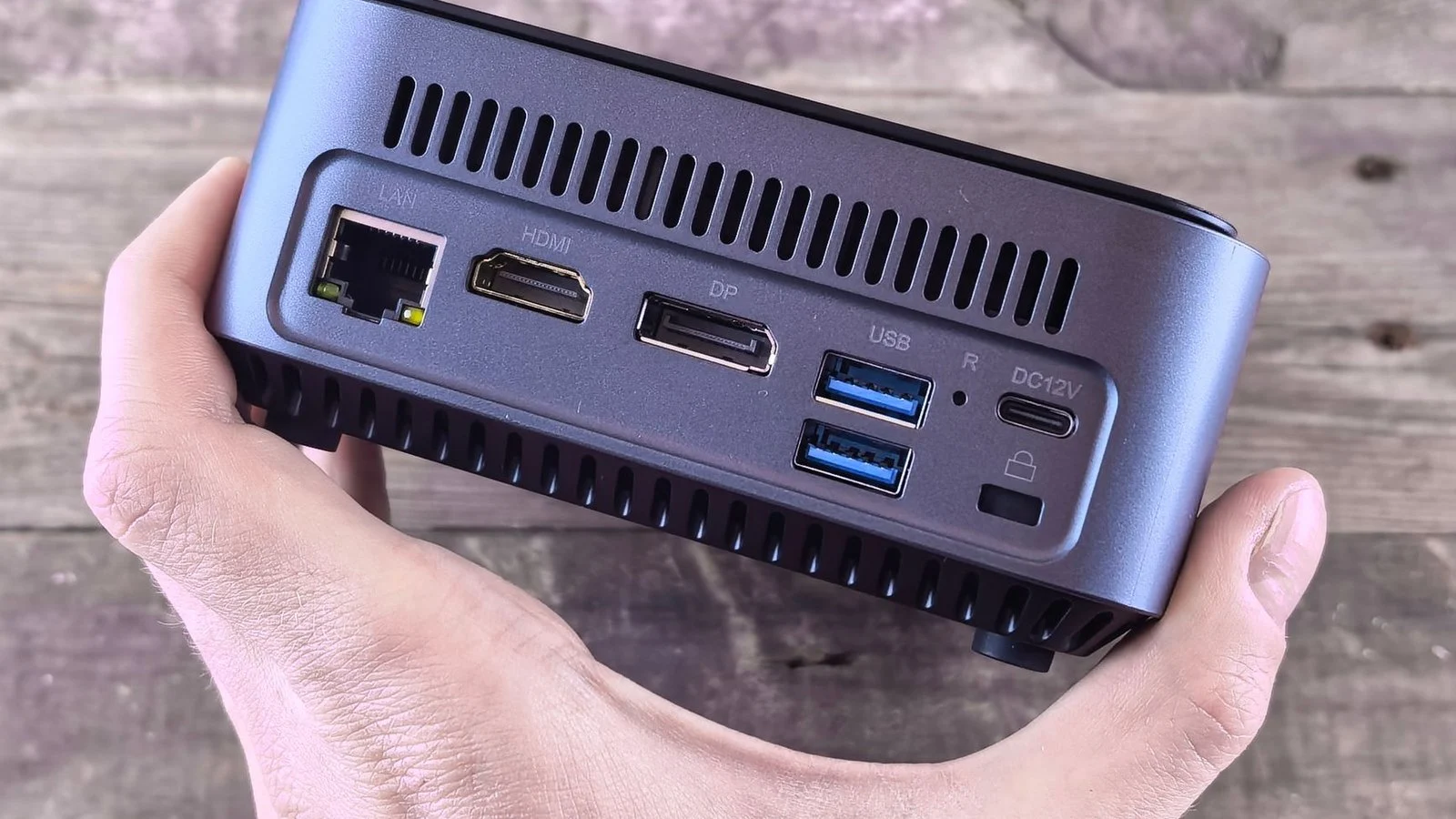 Hand holding a small gray mini PC showing LAN, HDMI, DP, USB, and power ports