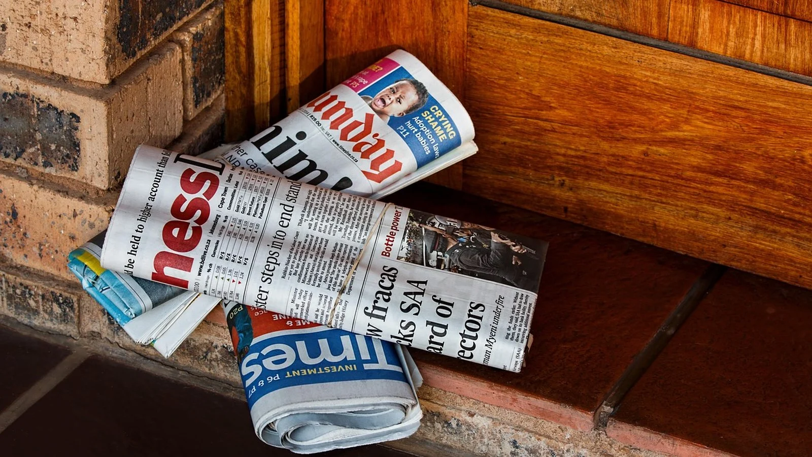 Rolled newspapers, including Sunday Times and Business Times, stacked on a wooden doorstep