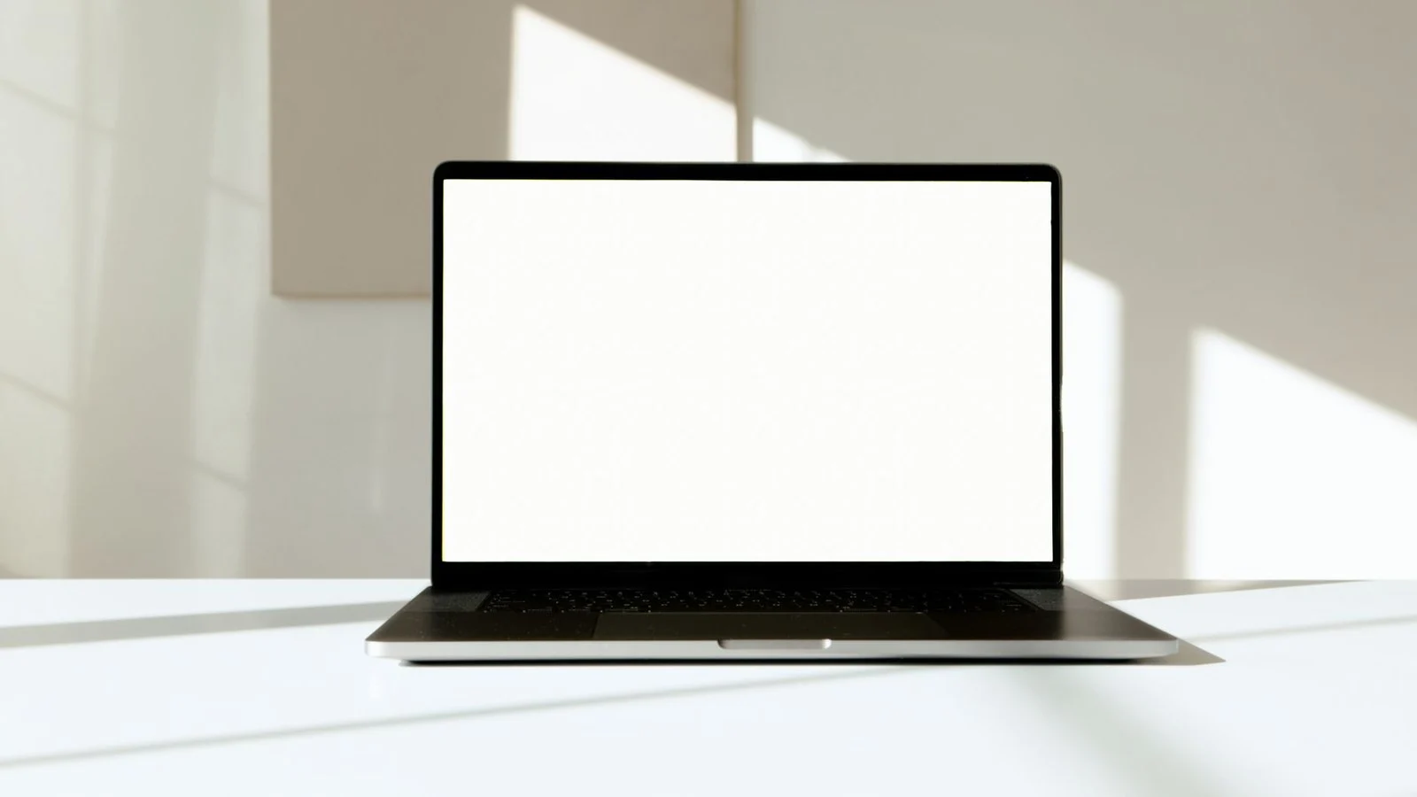 Silver laptop open on white desk with blank white screen and dark keyboard